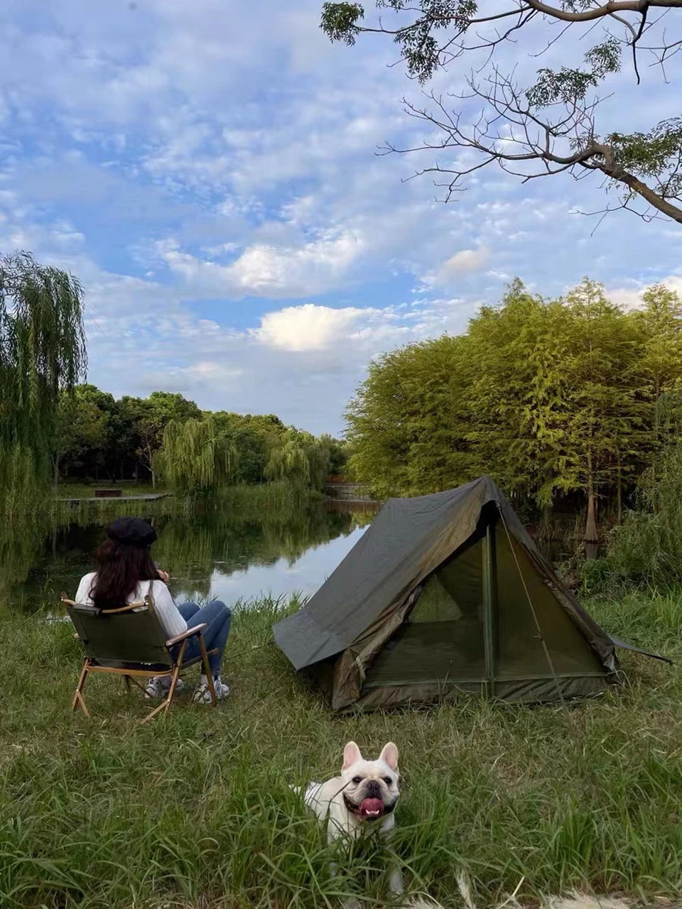 Green military camping tent pitched by a lakeside with camper in chair and dog in foreground