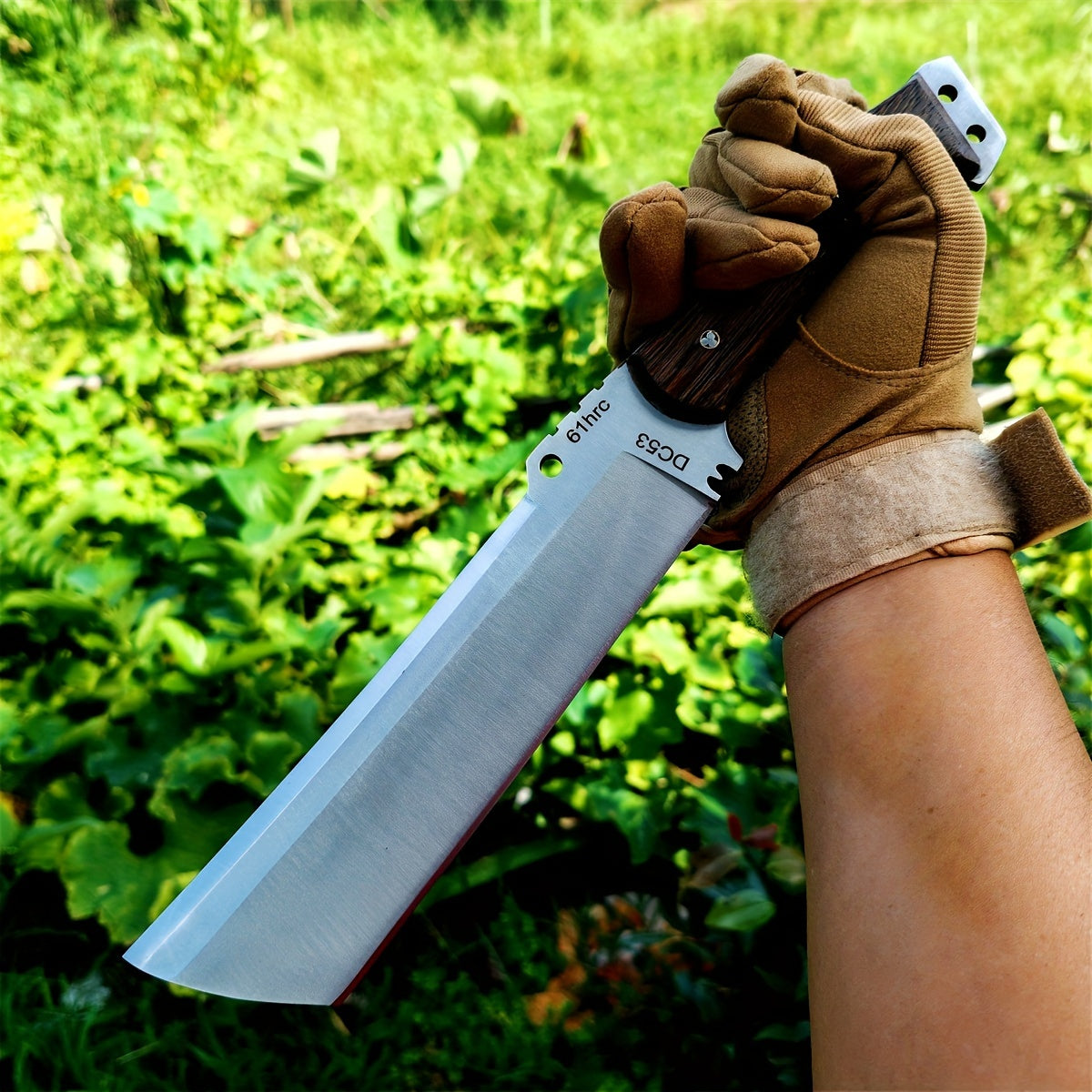 Gloved hand holding large cleaver style survival knife with wooden handle against green outdoor foliage