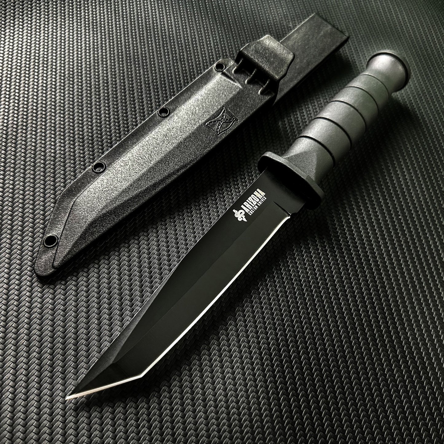 Black stainless steel army knife with textured grip and protective black sheath on carbon fiber background