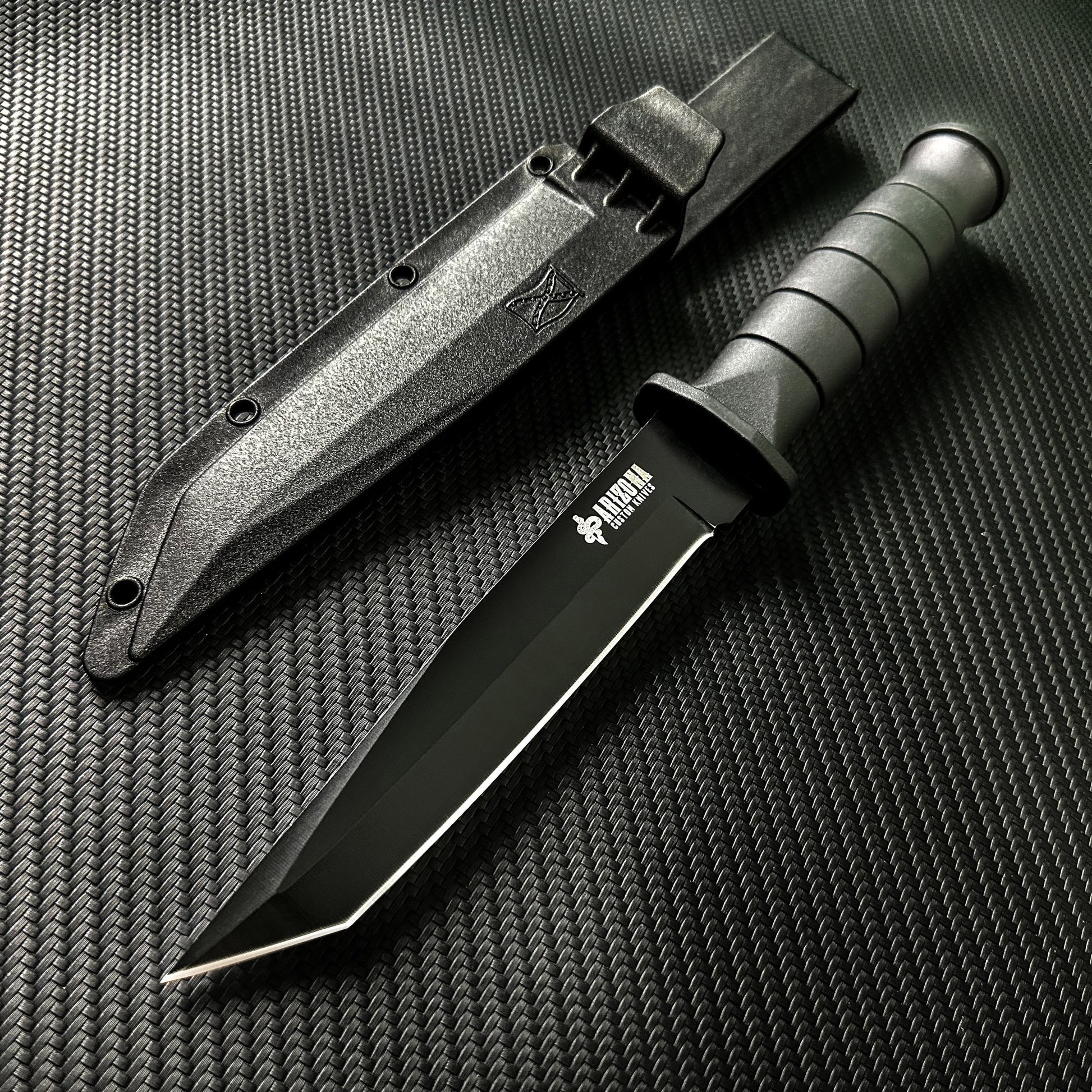 Black stainless steel army knife with textured grip and protective black sheath on carbon fiber background